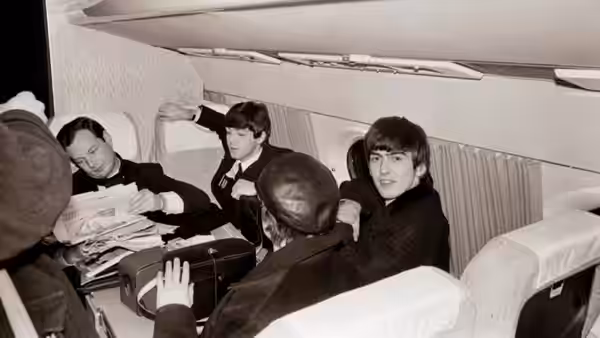 Three Beatles sit in an airplane cabin among luggage and newspapers