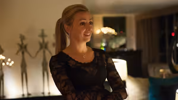 Lyndsey (Lisa McGrillis) stands smiling in a warmly lit living room