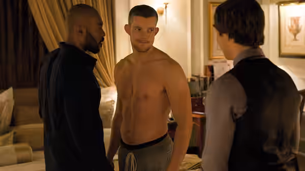 Ade and a shirtless man stand in a hotel room as another man faces them