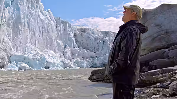 Al Gore stands beside a rocky shore, looking up at a towering glacier