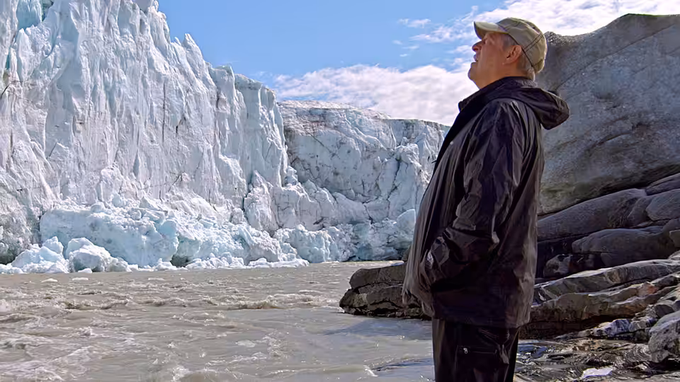 Al Gore stands beside a rocky shore, looking up at a towering glacier