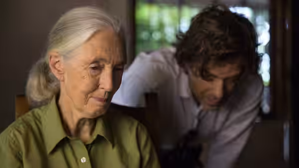 Jane Goodall sits thoughtfully as a man leans beside her indoors