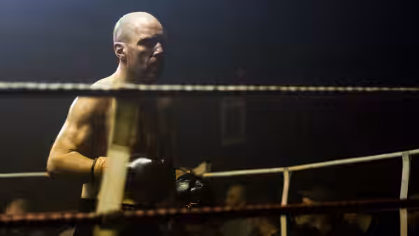 A bald boxer waits inside the ring, framed by ropes and ringside spectators