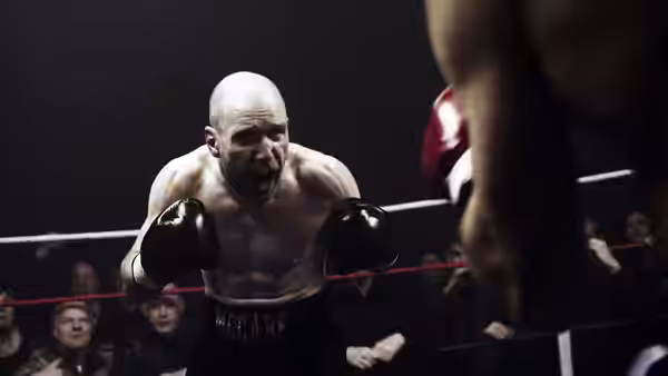 A bald boxer advances between the ropes as spectators watch from ringside