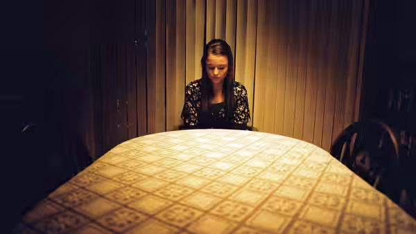 A young woman sits alone at a table in a wood-paneled room