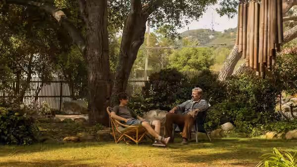 David and Nic sit facing each other in lawn chairs beneath a backyard tree
