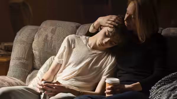 Karen comforts Nic on a living room couch, resting a hand on his head