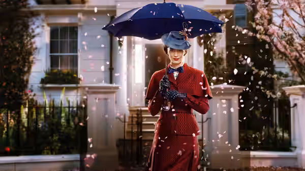 Mary Poppins (Emily Blunt) stands beneath her umbrella outside a London townhouse as petals swirl around her