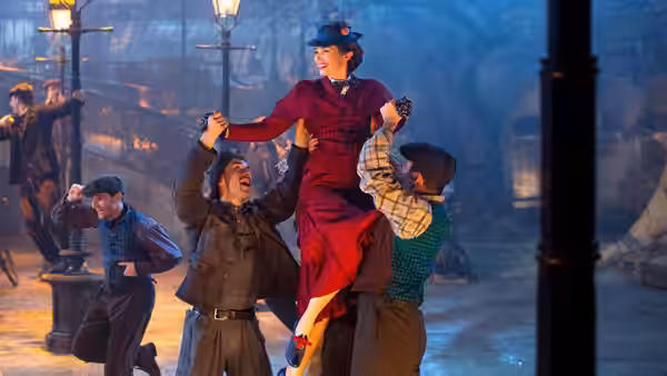 Mary Poppins (Emily Blunt) is lifted by cheering street sweepers on a lamplit London street
