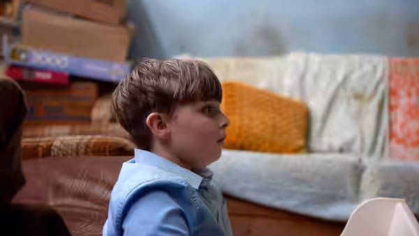 A young boy sits in a cluttered living room, looking to the side