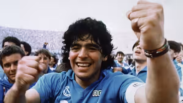 Diego Maradona celebrates in Napoli's blue kit, raising both fists among cheering fans
