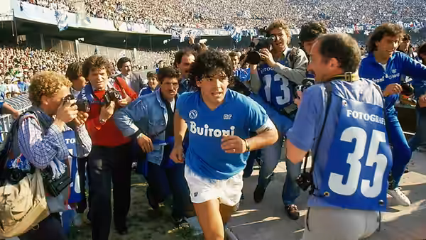 Diego Maradona runs through photographers in Napoli's blue kit as fans fill the stands