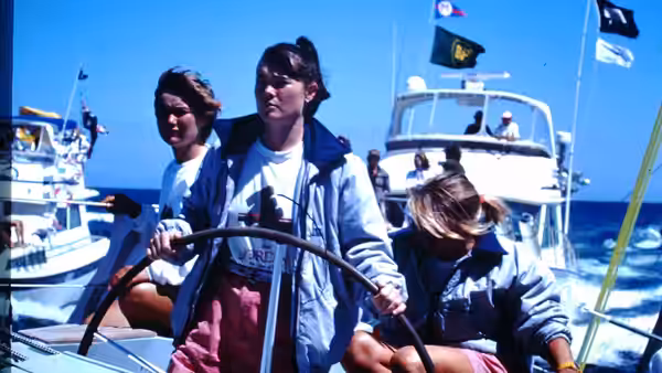 A woman steers a yacht among boats in a marina while two crewmates sit nearby