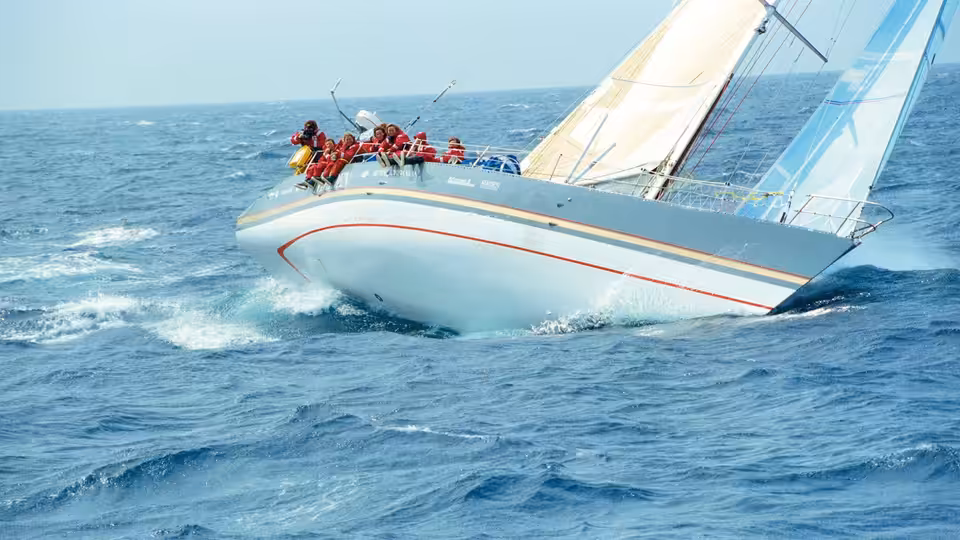 A crew of sailors leans over a yacht’s rail as it cuts through choppy water