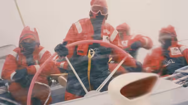 Sailors in red foul-weather gear steer a yacht through mist and spray