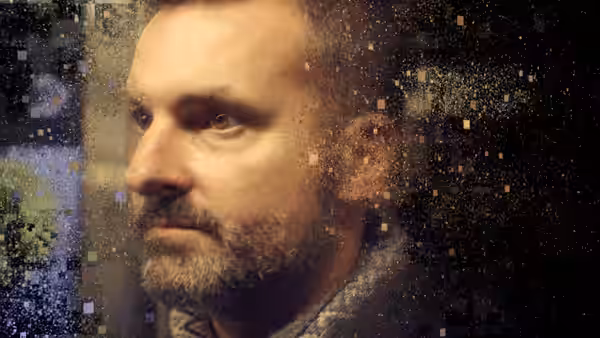 A bearded man’s face emerges among scattered digital squares