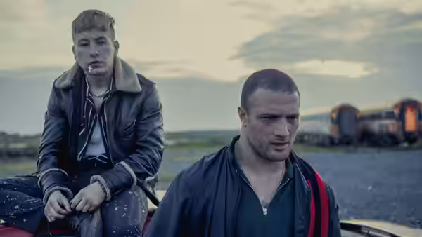 Dympna (Barry Keoghan) sits on a low wall while Arm (Cosmo Jarvis) stands beside railway tracks