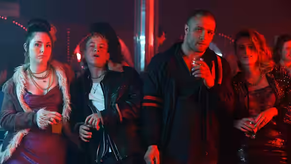Arm (Cosmo Jarvis) and Dympna (Barry Keoghan) stand with women in a red-lit nightclub