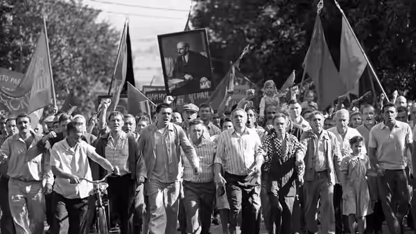 A crowd marches beneath flags and a large portrait, filling a tree-lined street