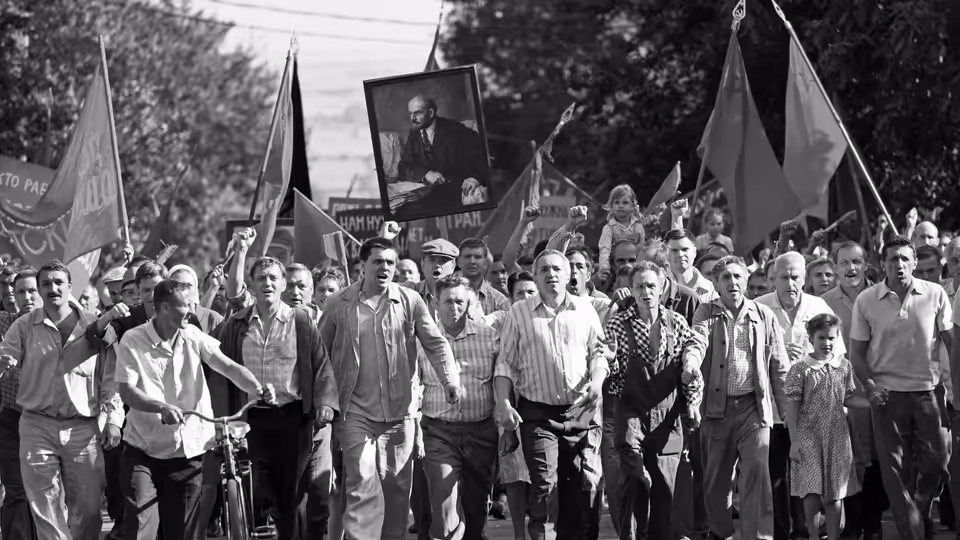 A crowd marches beneath flags and a large portrait, filling a tree-lined street