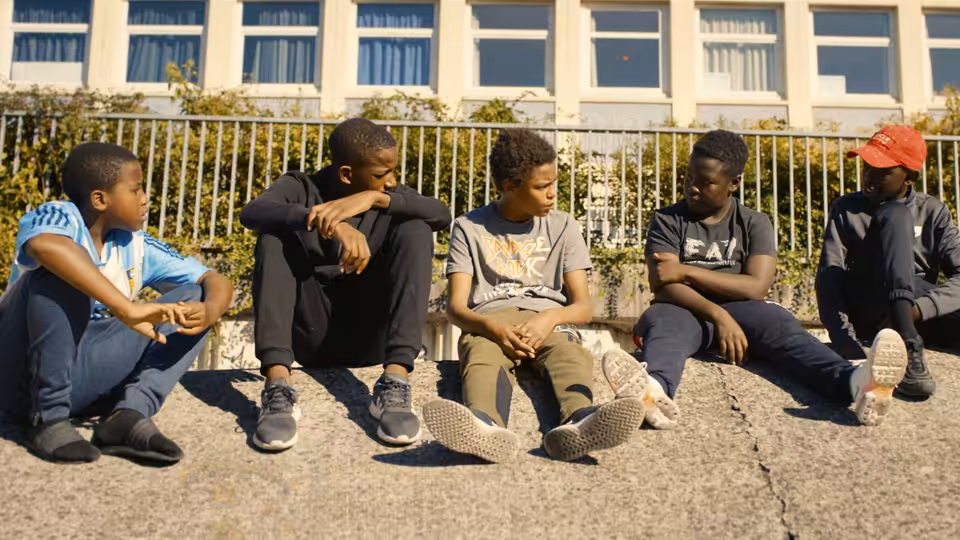Five boys sit together on a sunny sidewalk outside an apartment building