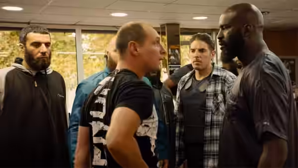 A bald, bearded man faces off with several men inside a room