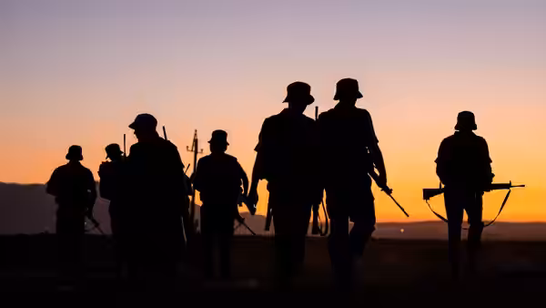 Silhouetted soldiers carrying rifles walk across open ground at sunset