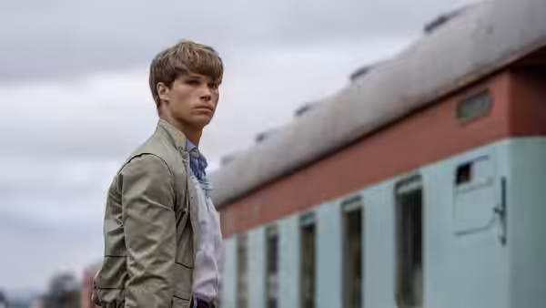 A young man stands beside a passenger train under a cloudy sky