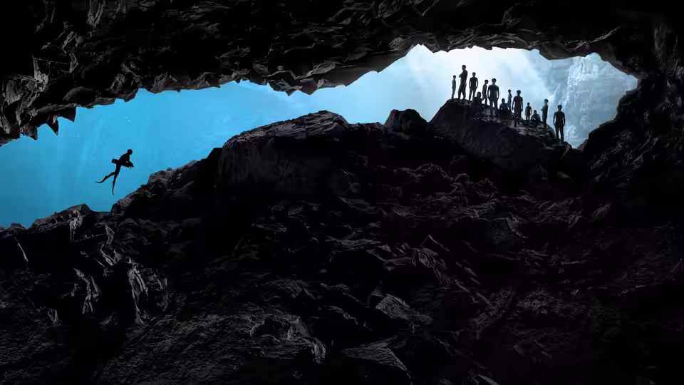 A diver jumps from rocks into blue water while a group watches from a cave ledge