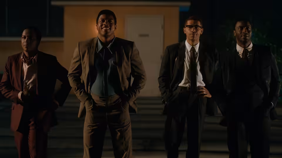 Four men in suits stand together outside a building at night