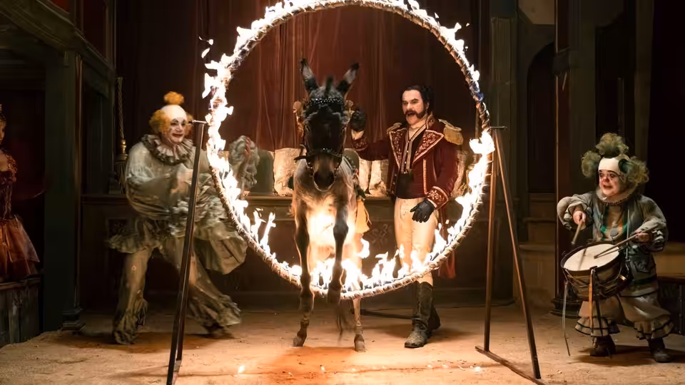 Mangiafuoco stands beside a donkey passing through a ring of fire, with two performers nearby