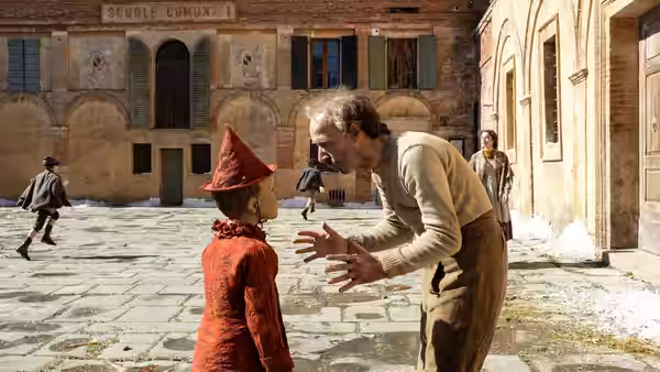 Mister Geppetto gestures to Pinocchio in a town square outside a school