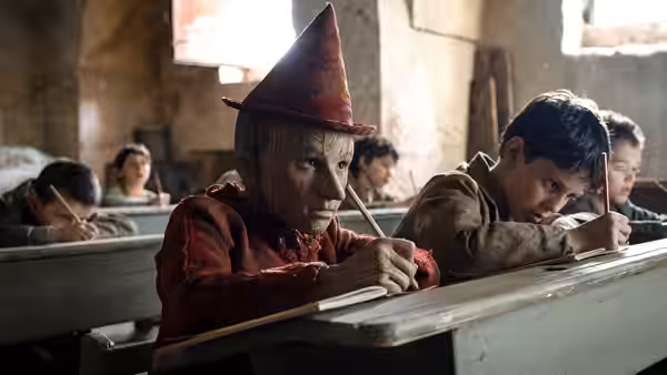 Pinocchio writes at a school desk beside a boy as classmates study
