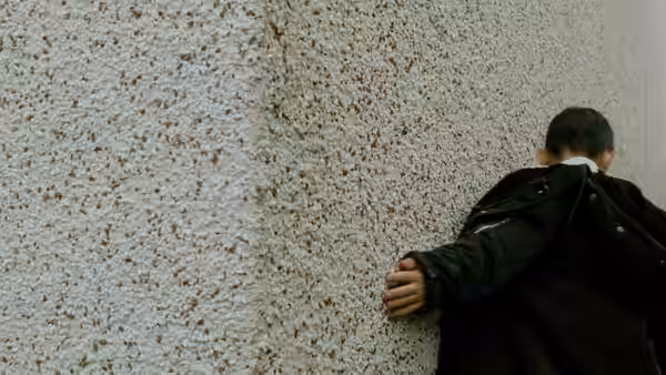 A boy in a black jacket leans against a white textured wall