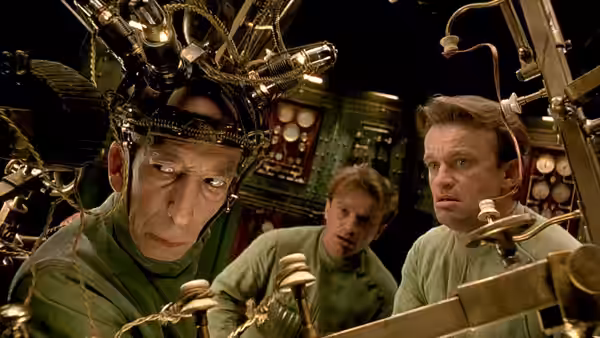 A man in a metal head harness works at controls while two men watch