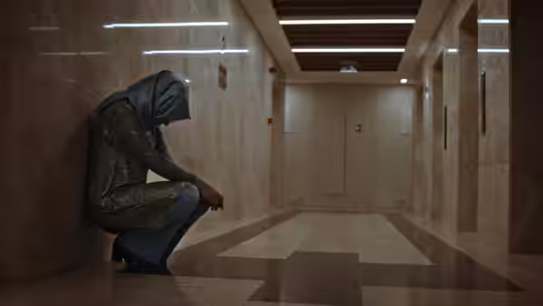A hooded person crouches against a marble wall in an empty corridor