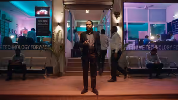 Balram (Adarsh Gourav) stands outside a technology company entrance as people pass behind him