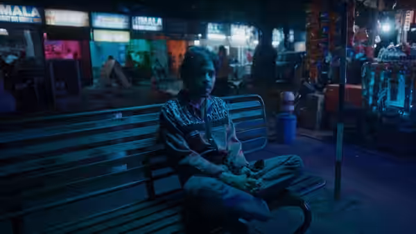 Balram (Adarsh Gourav) sits cross-legged on a bench in a neon-lit market