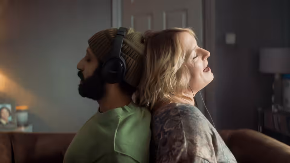 Ali (Adeel Akhtar) and Ava (Claire Rushbrook) sit back to back, sharing headphones indoors