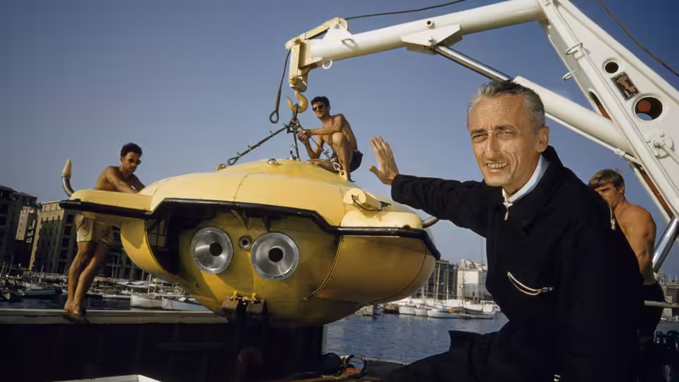 Jacques Cousteau beside a yellow research submersible being lifted aboard a vessel