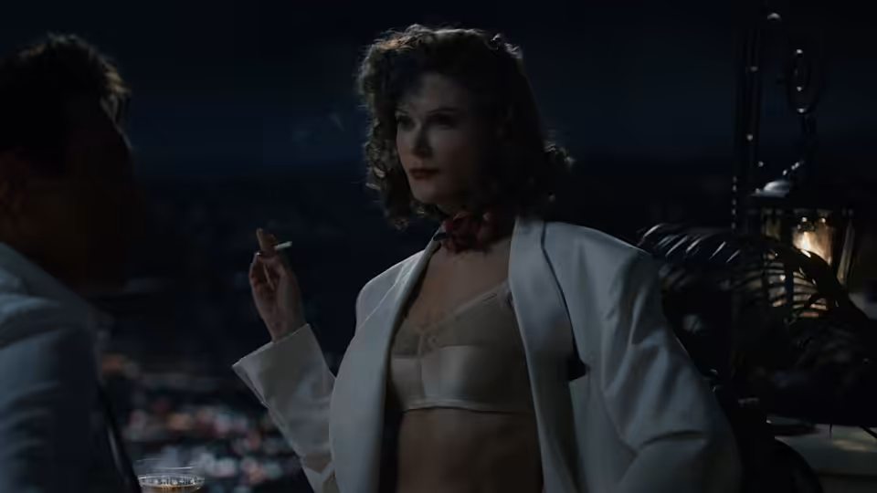 A woman in a white robe holds a cigarette beside a man in a dim room