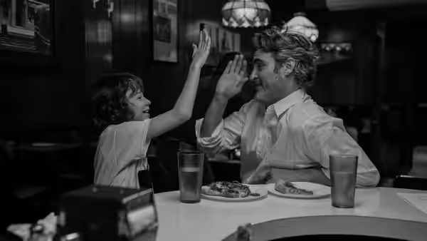 Johnny (Joaquin Phoenix) and Jesse (Woody Norman) raise their hands across a restaurant table
