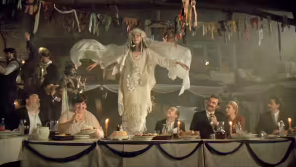 a bride in white standing on a table with arms outstretched at a feast