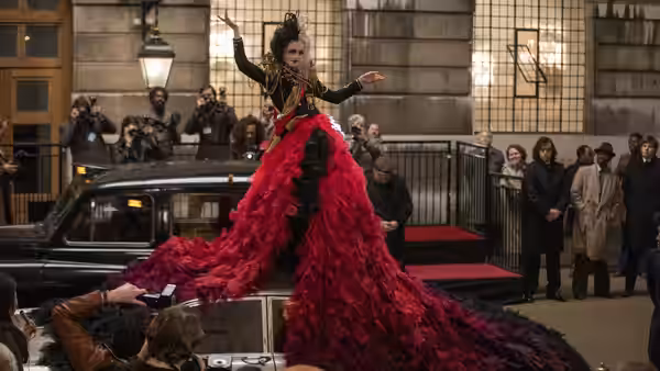 Cruella (Emma Stone) stands atop a car in a sweeping red gown before photographers