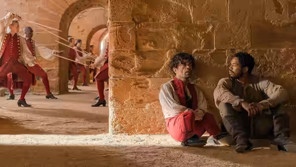Cyrano (Peter Dinklage) and Christian (Kelvin Harrison Jr.) rest against a stone wall as soldiers fence nearby