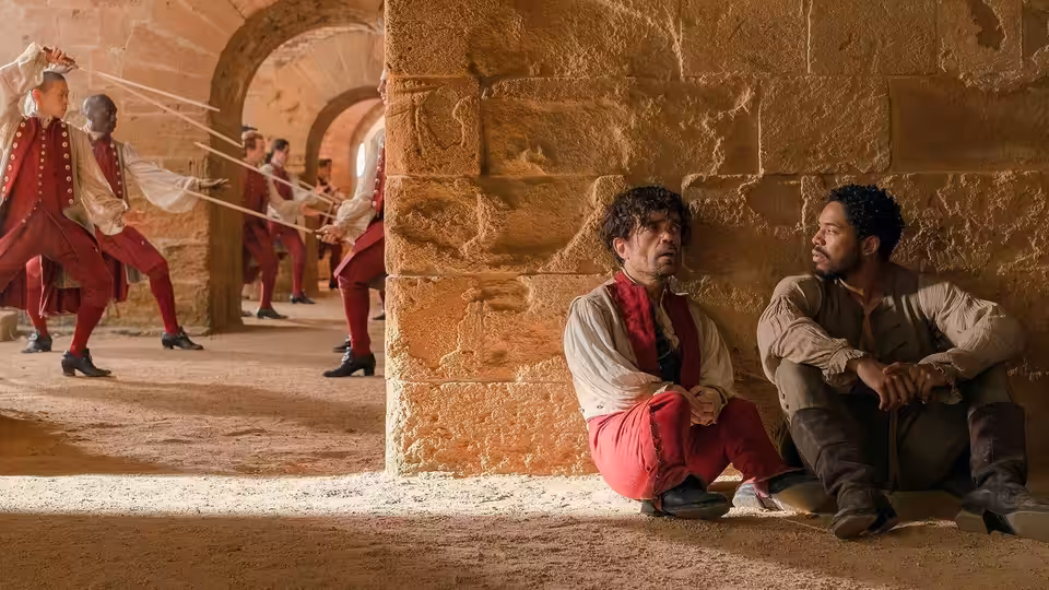 Cyrano (Peter Dinklage) and Christian (Kelvin Harrison Jr.) rest against a stone wall as soldiers fence nearby
