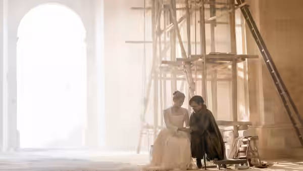 A woman and man sit together beneath scaffolding inside a bright stone building