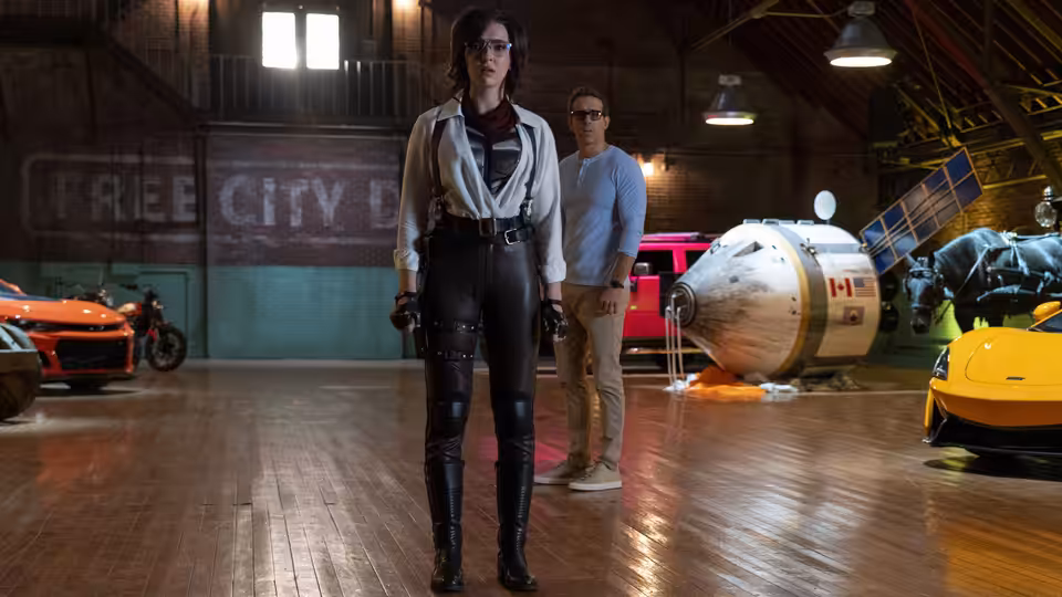 Millie (Jodie Comer) stands in a workshop as Guy (Ryan Reynolds) looks on behind her