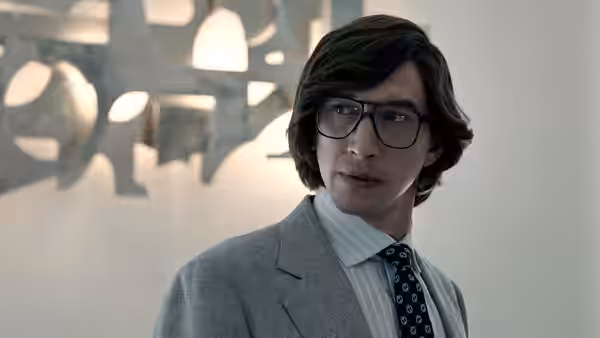 Maurizio Gucci (Adam Driver) stands in an office wearing a gray suit and glasses