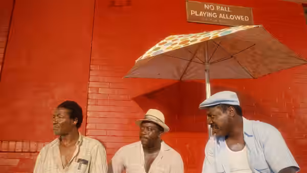 Three men sit on the sidewalk against a red wall under an umbrella